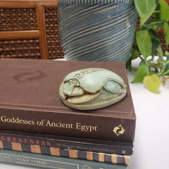 Decorative scarab object on a book titled 'Goddesses of Ancient Egypt' with plants and wooden elements in the background.