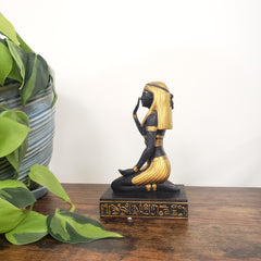 Decorative Egyptian statue with black and gold design on a wooden post, surrounded by green leaves.