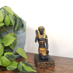 Decorative statue of a figure with gold accents on a wooden base, surrounded by green plants.