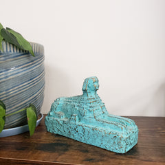 Turquoise Egyptian Sphinx Statue on a wooden surface with a striped pillow and green leaves in the foreground.