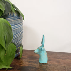 Turquoise Egyptian goddess Hathor bust statue on a wooden surface with green leaves in the foreground.