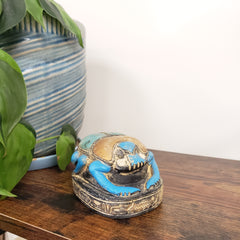 Decorative blue and gold Egyptian scarab statue on a wooden surface with plants in the foreground.