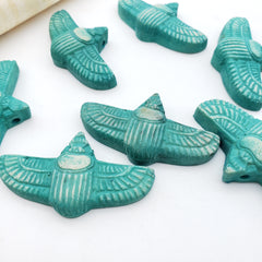 Turquoise scarab-shaped beads with carved feather details, arranged on a white surface.