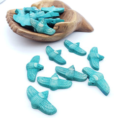 Turquoise scarab-shaped beads with carved feather patterns, shown scattered on a white background with a wooden bowl containing more of the same.