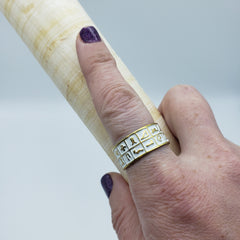 Hand wearing a gold ring with symbols holding a wooden object against a light background