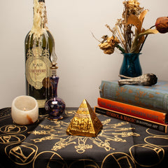 Decorative setup with books, a bottle, a pyramid, and a candle on a tablecloth with symbols.
