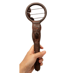 A hand holding a dark brown carved sistrum with an Egyptian god head design on the handle and a circular loop at the end with metal blades.