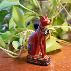 Red cat figurine with hieroglyphics on a wooden surface with green plants in the background