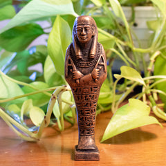 Bronze-colored Egyptian sphinx figurine on a wooden surface with green plants in the background