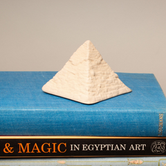 A small textured off-white pyramid figurine displayed on top of a blue book, with the spine '& MAGIC IN EGYPTIAN ART' visible at the bottom.