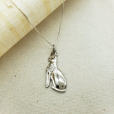 A silver pendant necklace featuring a cat design, with a chain and a crystal inlay on the pendant. The pendant is on a beige fabric background.