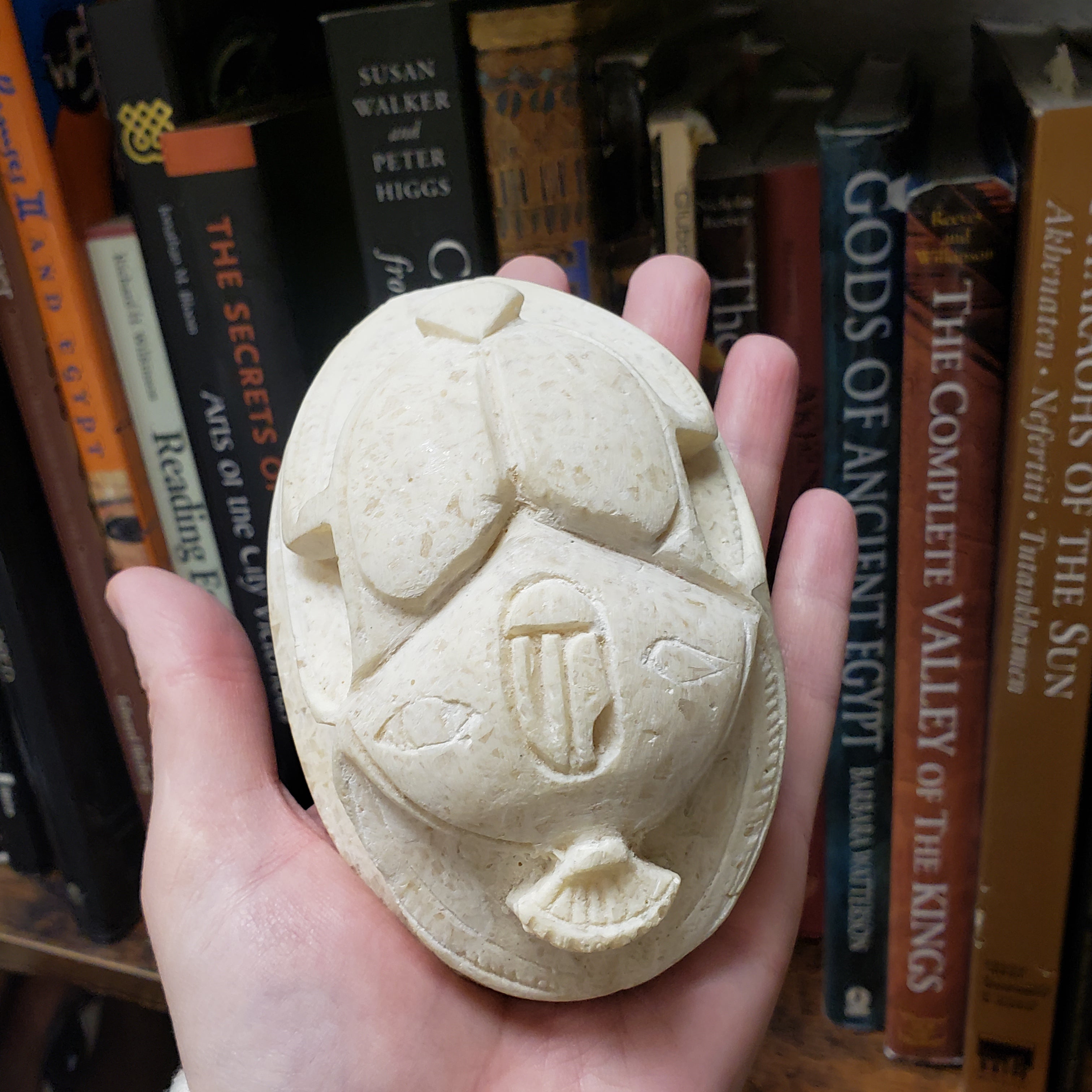 Hand holding a small stone sculpture of a face with hieroglyphs against a background of books on Egyptian history.