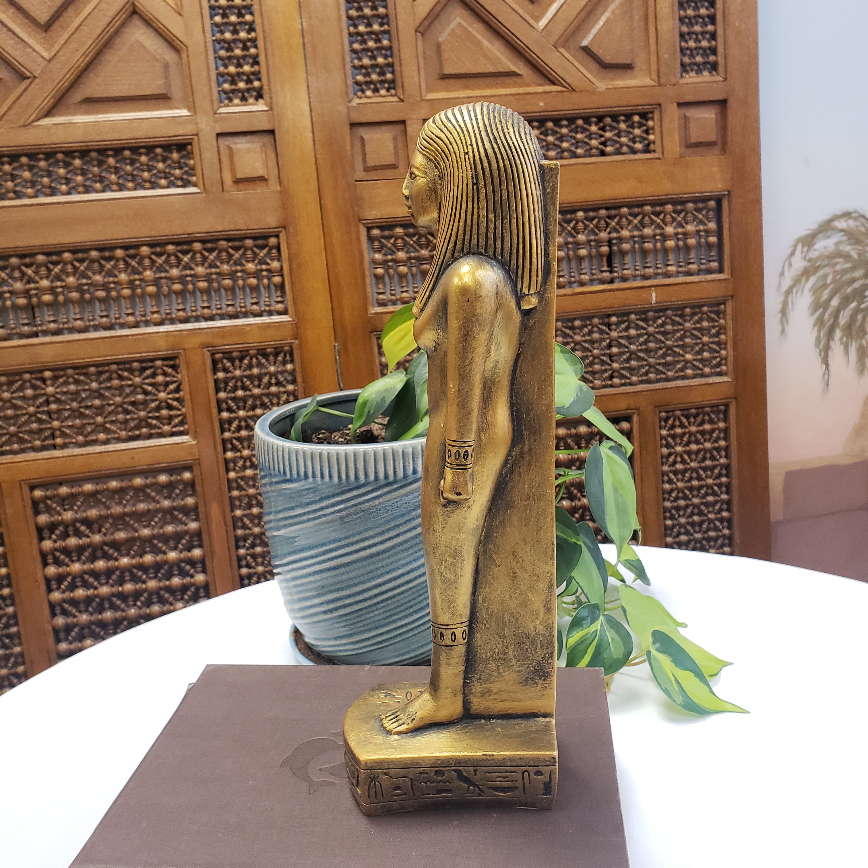 Decorative gold statue on a stand with a wooden panel and plant in the background