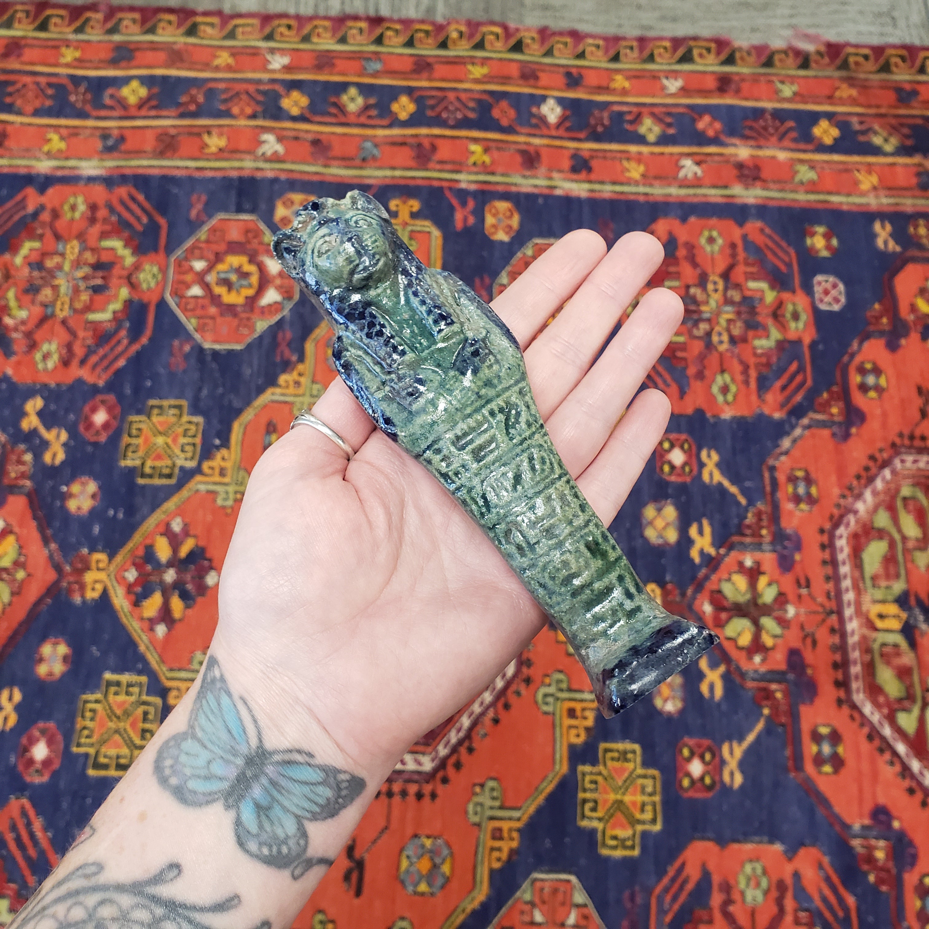 Hand holding a green Egyptian Sekhmet figure against a colorful rug background