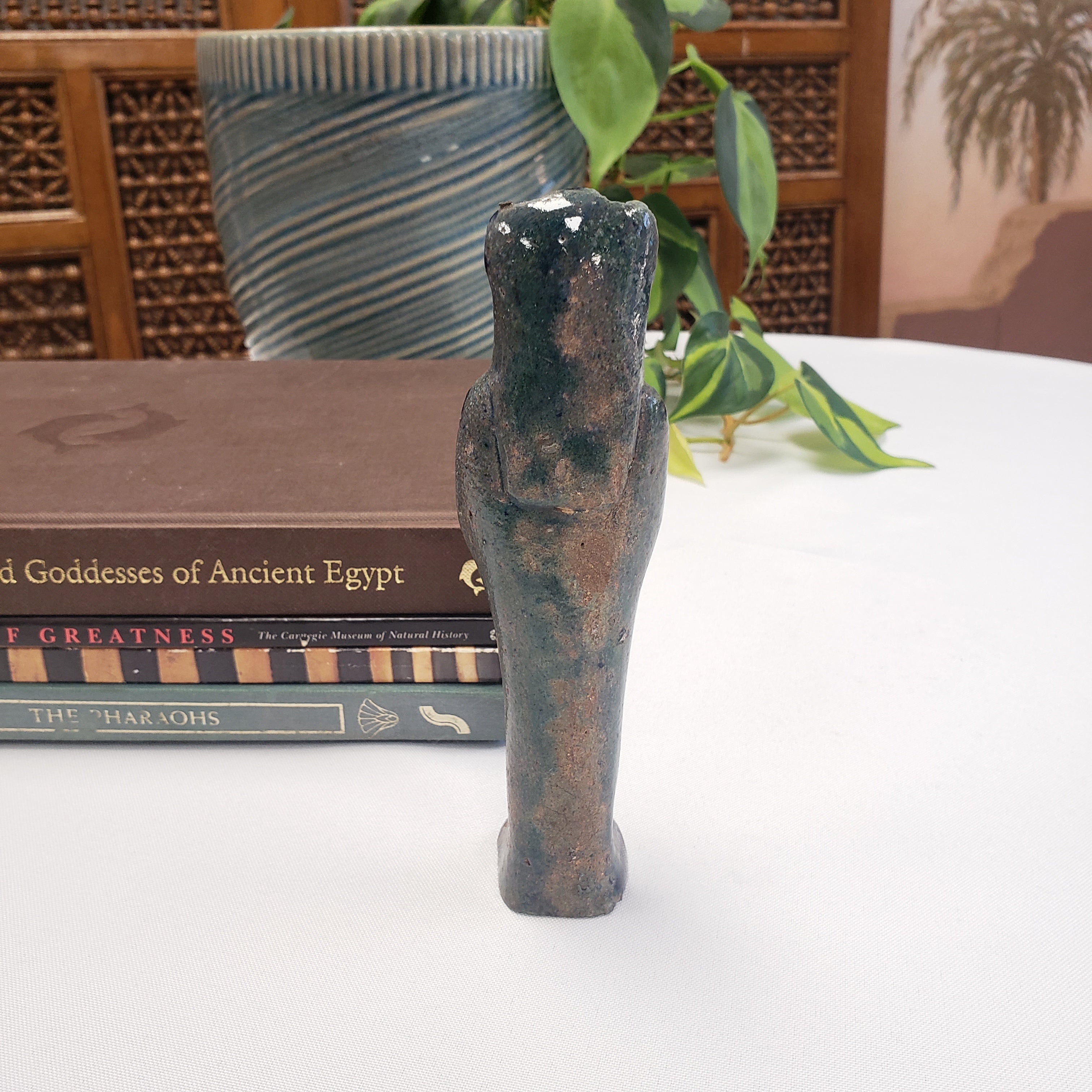 Decorative object resembling a ancient Egyptian Egyptian Sekhmet figure on top of books with a plant and decorative wall in the background.