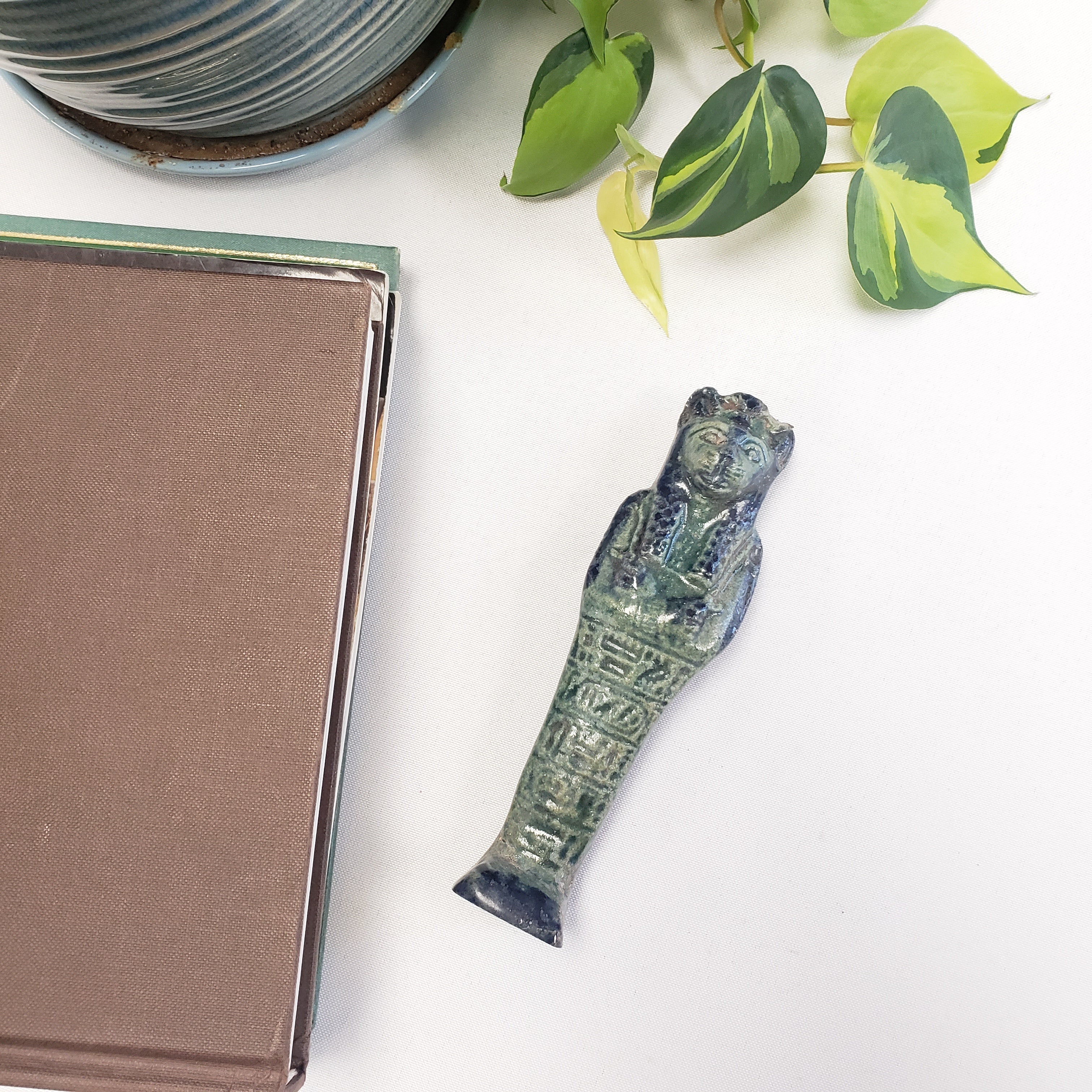 Decorative green ceramic Egyptian Sekhmet figure on a book with a plant in the corner