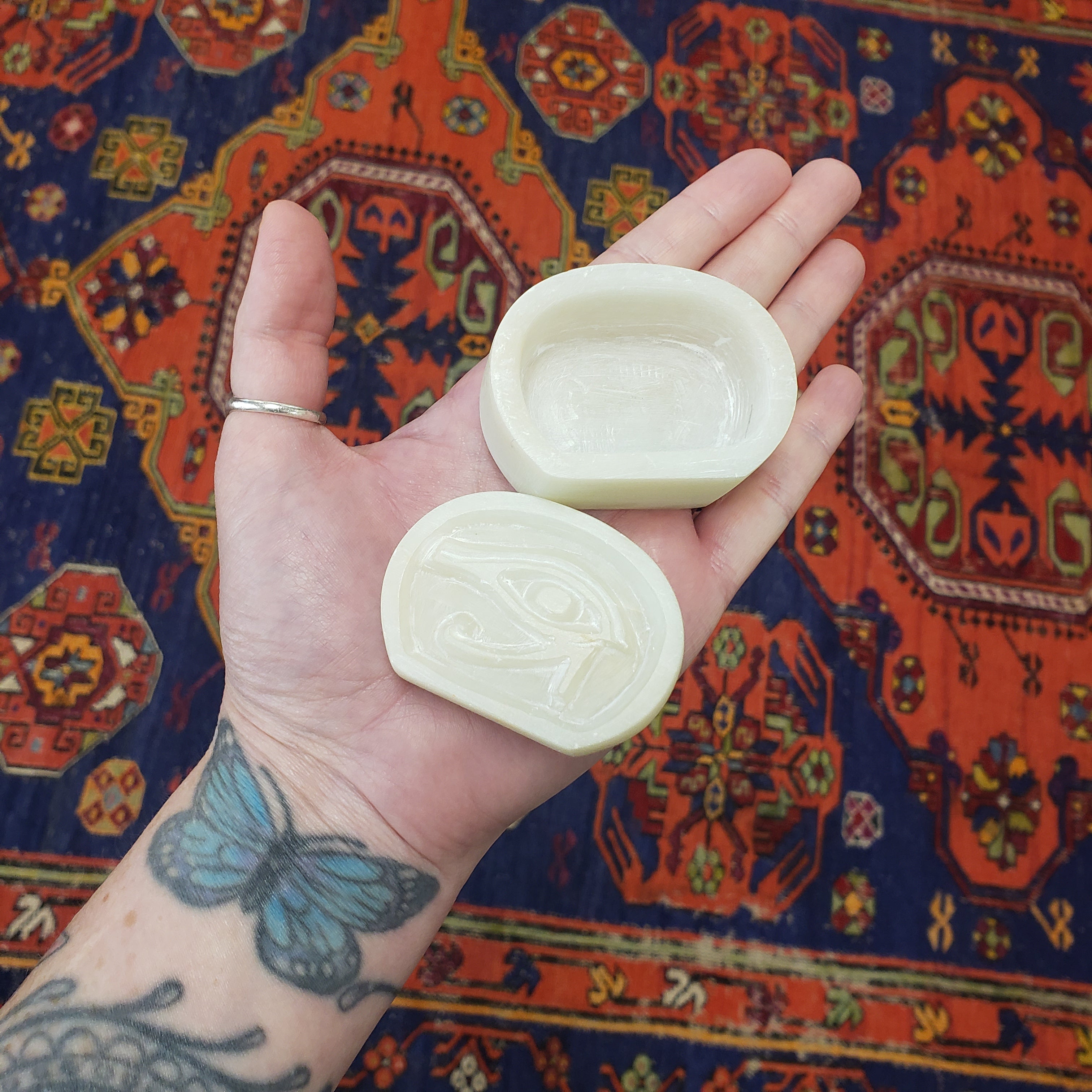 Hand holding White soapstone box with an eye symbol with intricate designs against a colorful patterned rug.