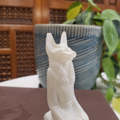 White marble sculpture of Anubis against a blurred indoor background with plants.