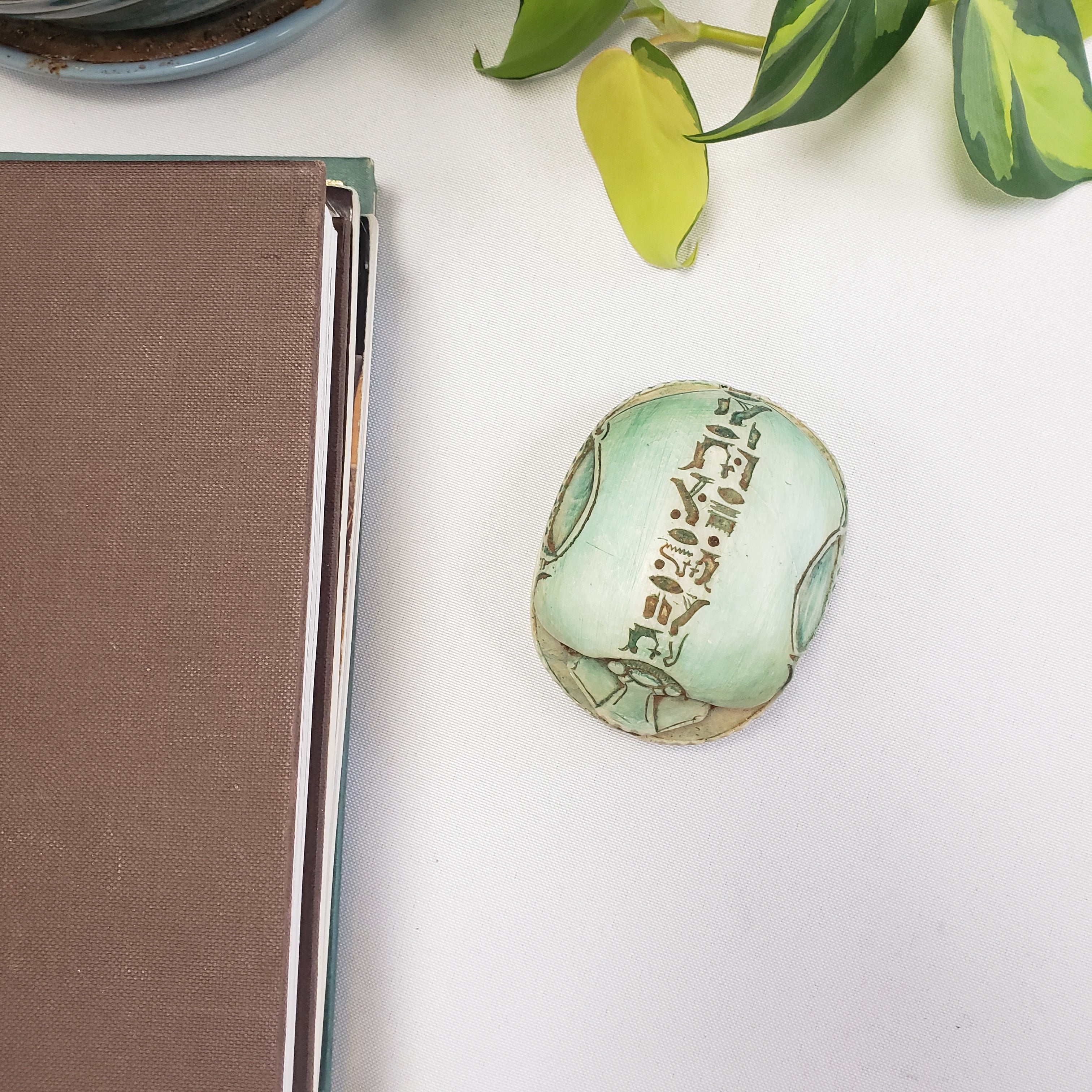 Green stone scarab with engraved text on a white surface next to a plant