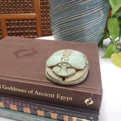 Decorative scarab object on a book titled 'Goddesses of Ancient Egypt' with a plant in the background.