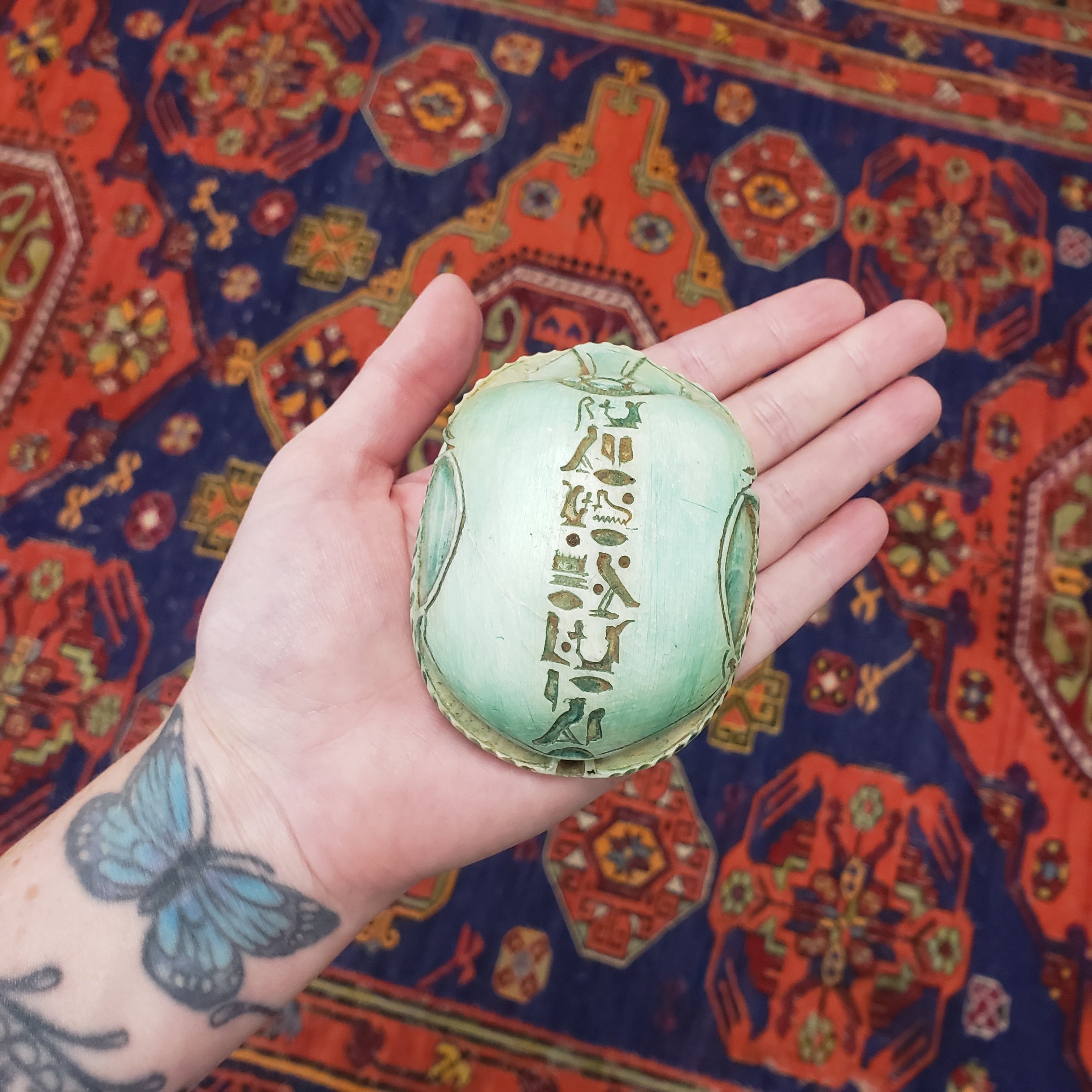 Hand holding a green decorative scarab item with intricate patterns against a colorful patterned fabric background