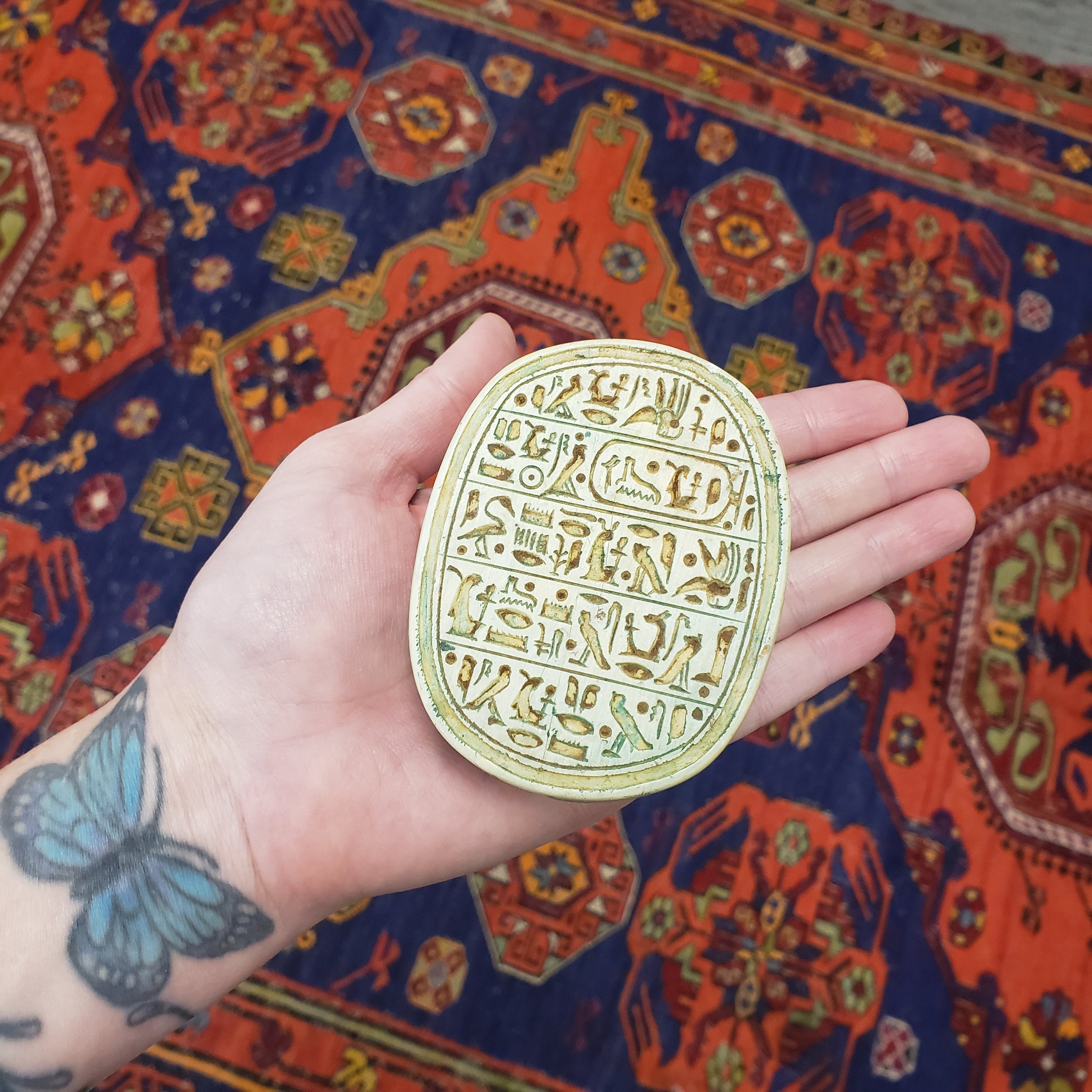 Hand holding a gold circular scarab object with engraved text against a colorful patterned rug.