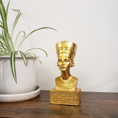 Gold bust of a historical figure on a wooden surface with a plant in the background