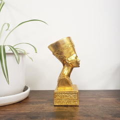 Gold bust of a historical figure on a wooden surface with a plant in the background