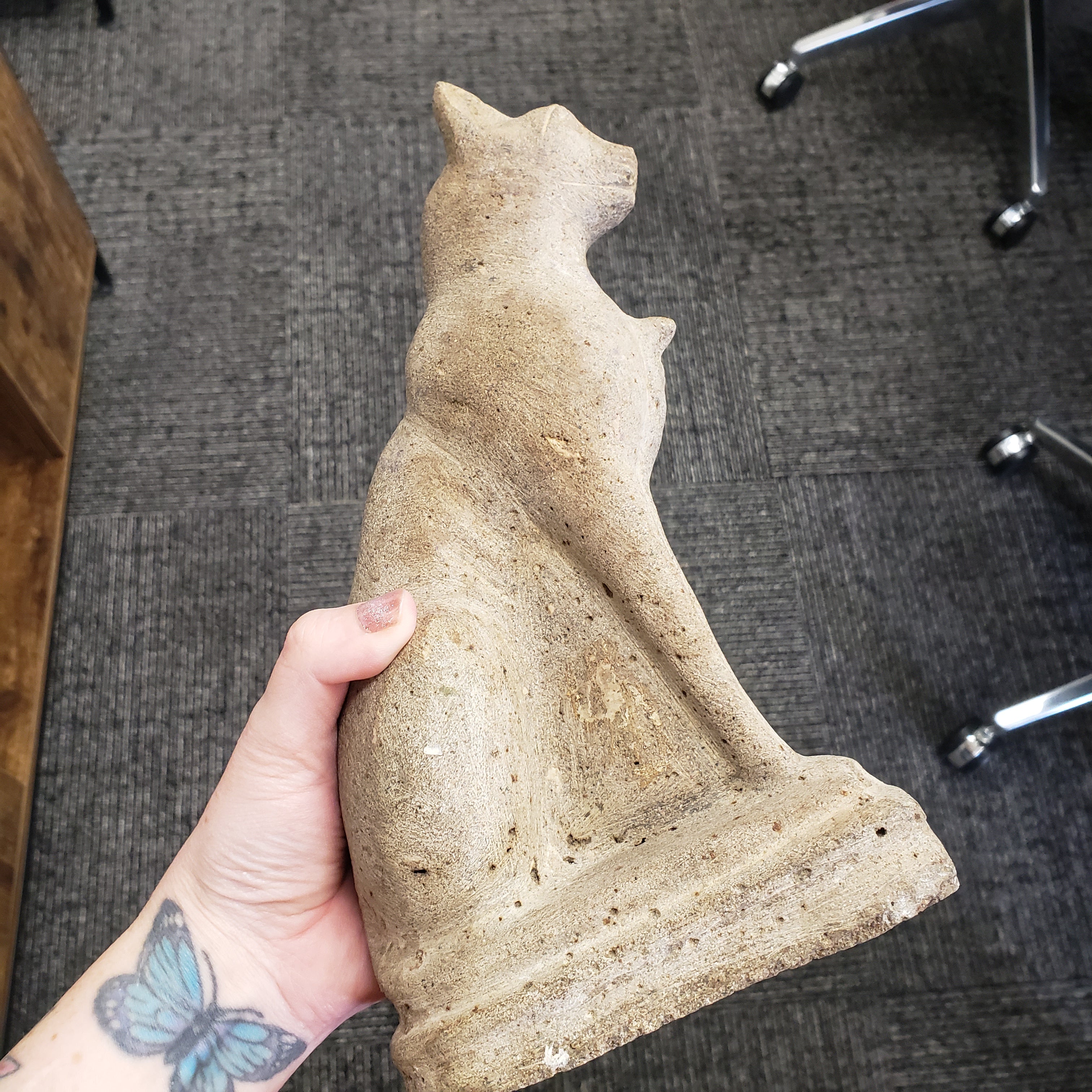Hand holding a stone sculpture of a cat on a gray carpeted floor.