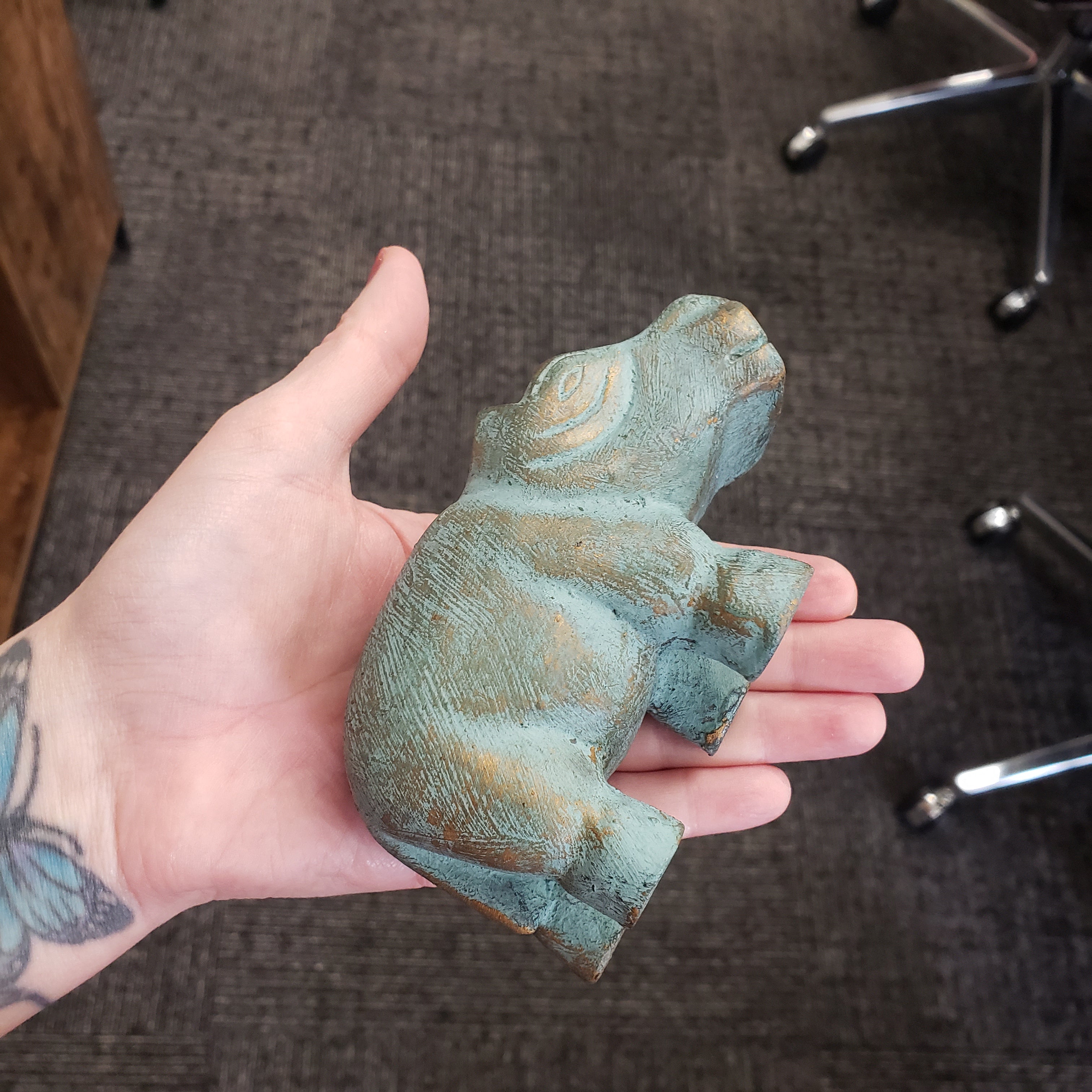 Hand holding a small, textured hippo statue object with a greenish-brown patina on a carpeted floor.