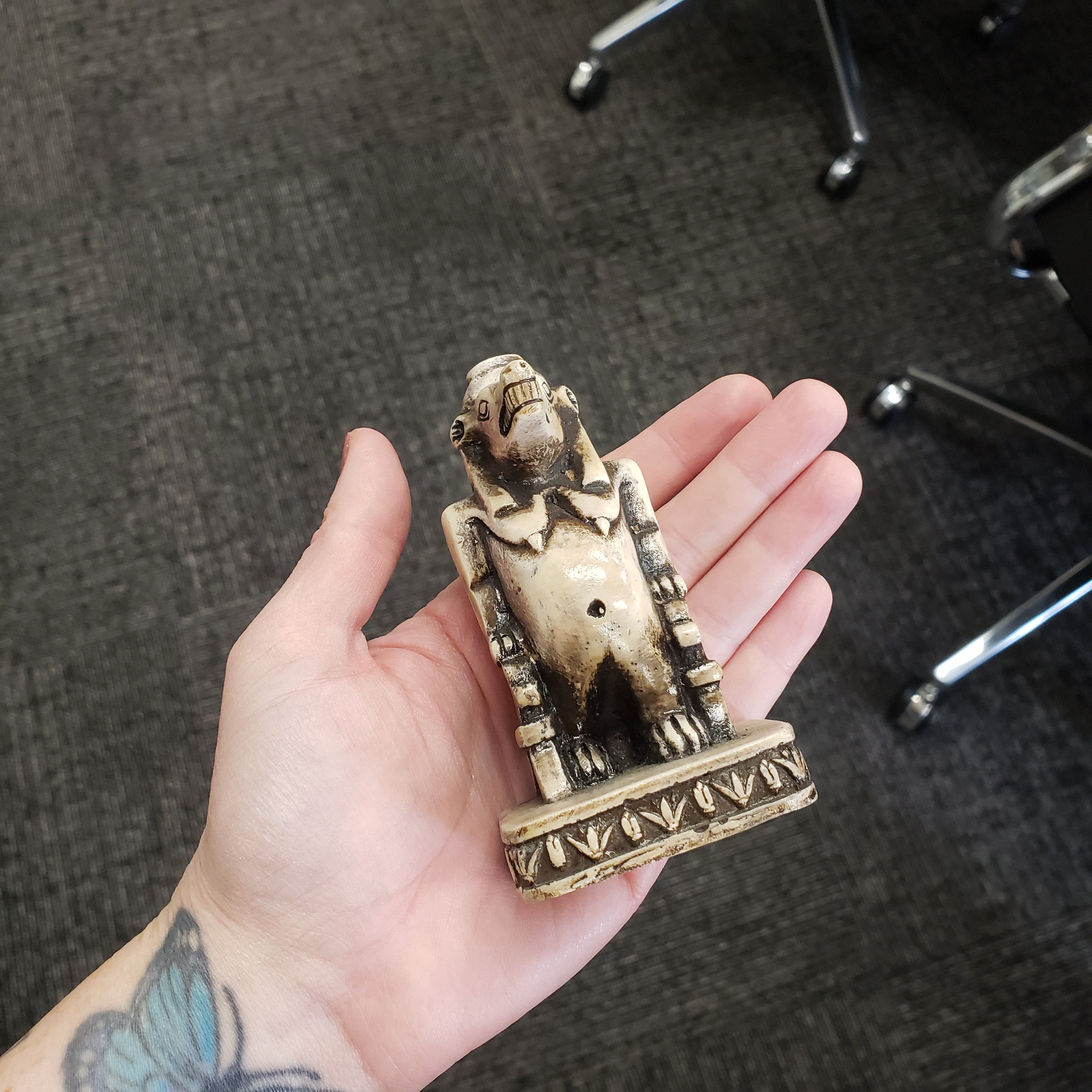 Hand holding a small ornate Egyptian hippo goddess object on a carpeted floor.