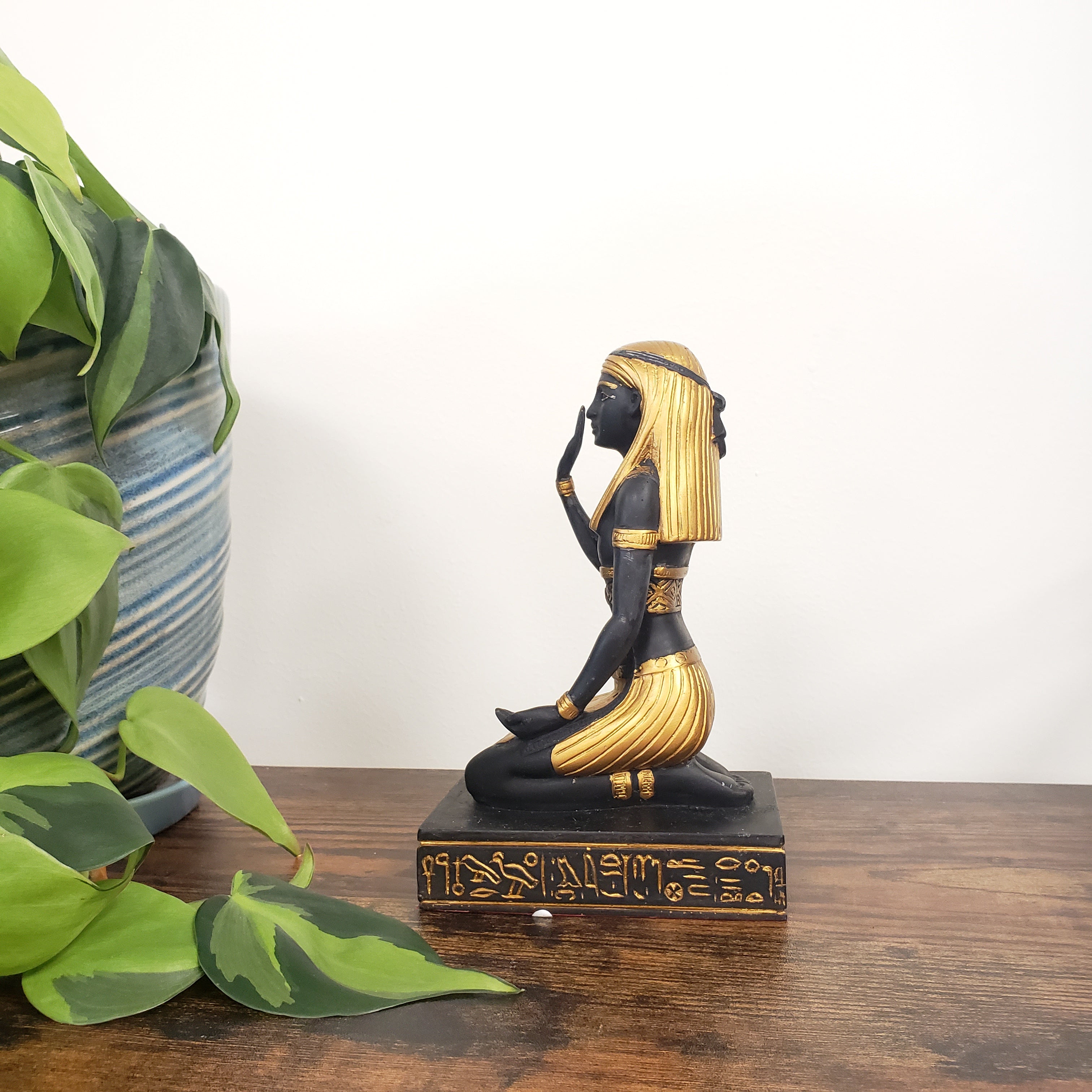 Decorative Egyptian statue with black and gold design on a wooden post, surrounded by green leaves.