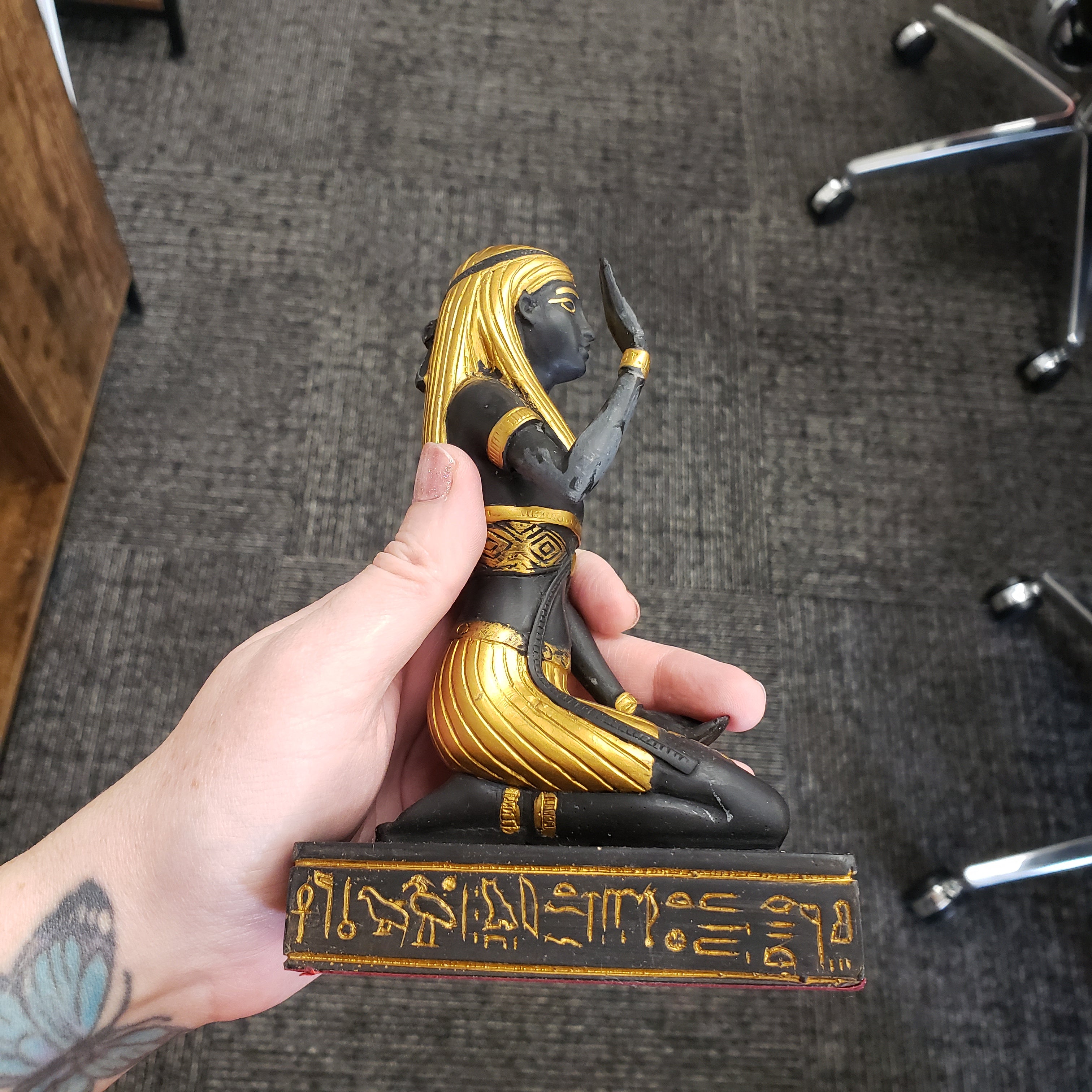 Hand holding a decorative Egyptian statue object with gold and black design, featuring hieroglyphics, on a carpeted floor.