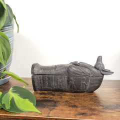 Decorative Anubis on a wooden panel with green leaves in the foreground.