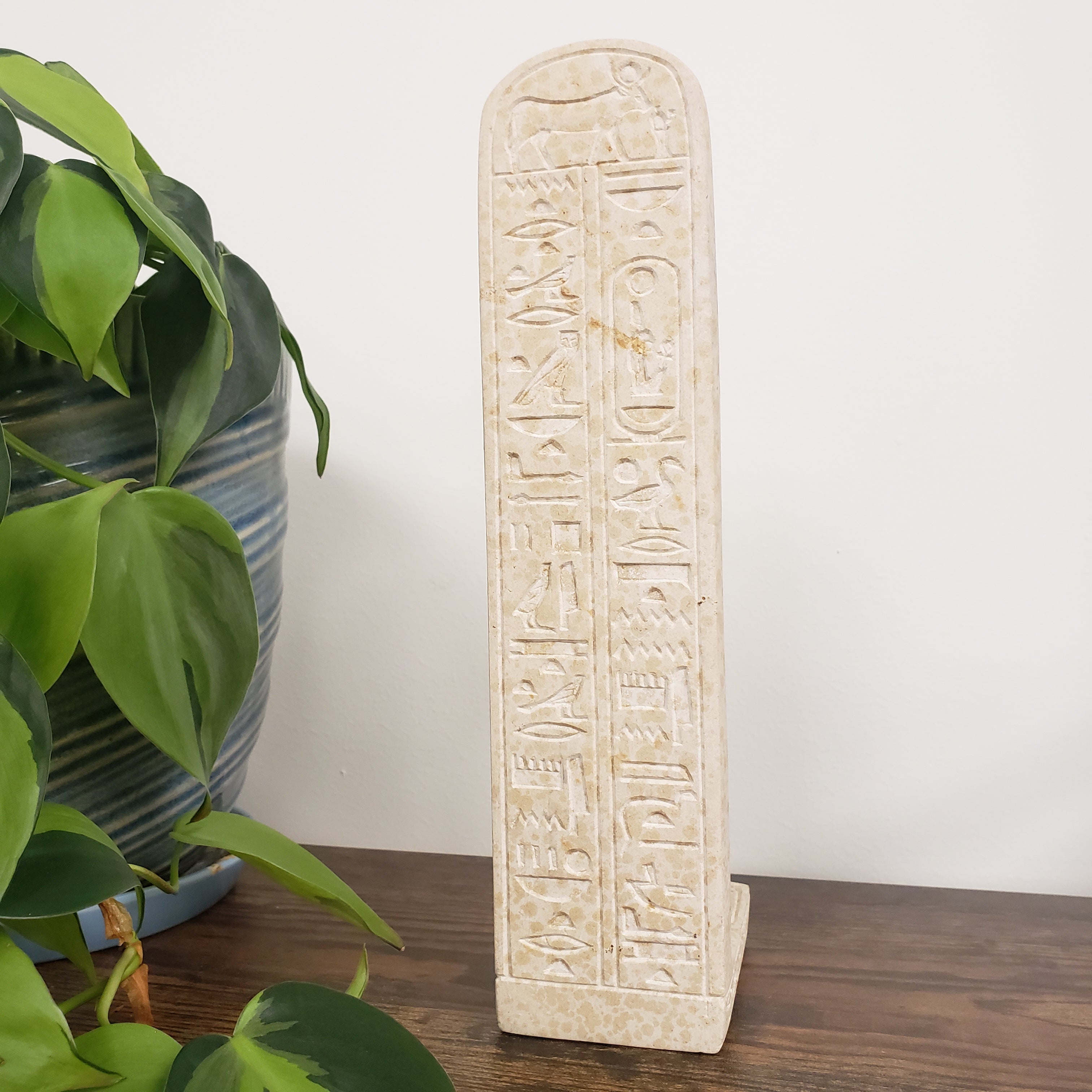 Decorative stone statue of Tiye with hieroglyphics on a white wall with green plants in the foreground