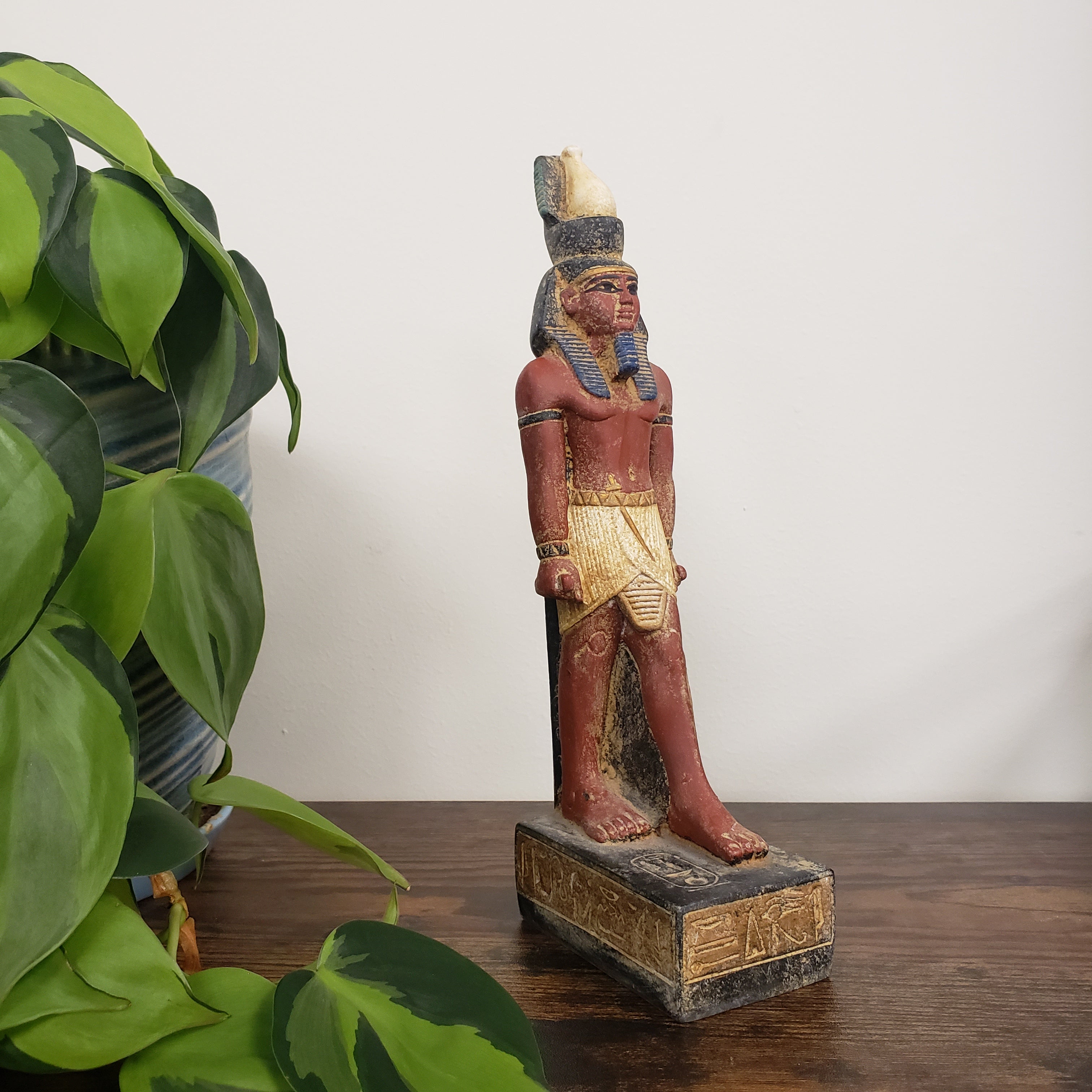 Decorative Egyptian Ramses statue mounted on a wooden panel with green leaves in the foreground.