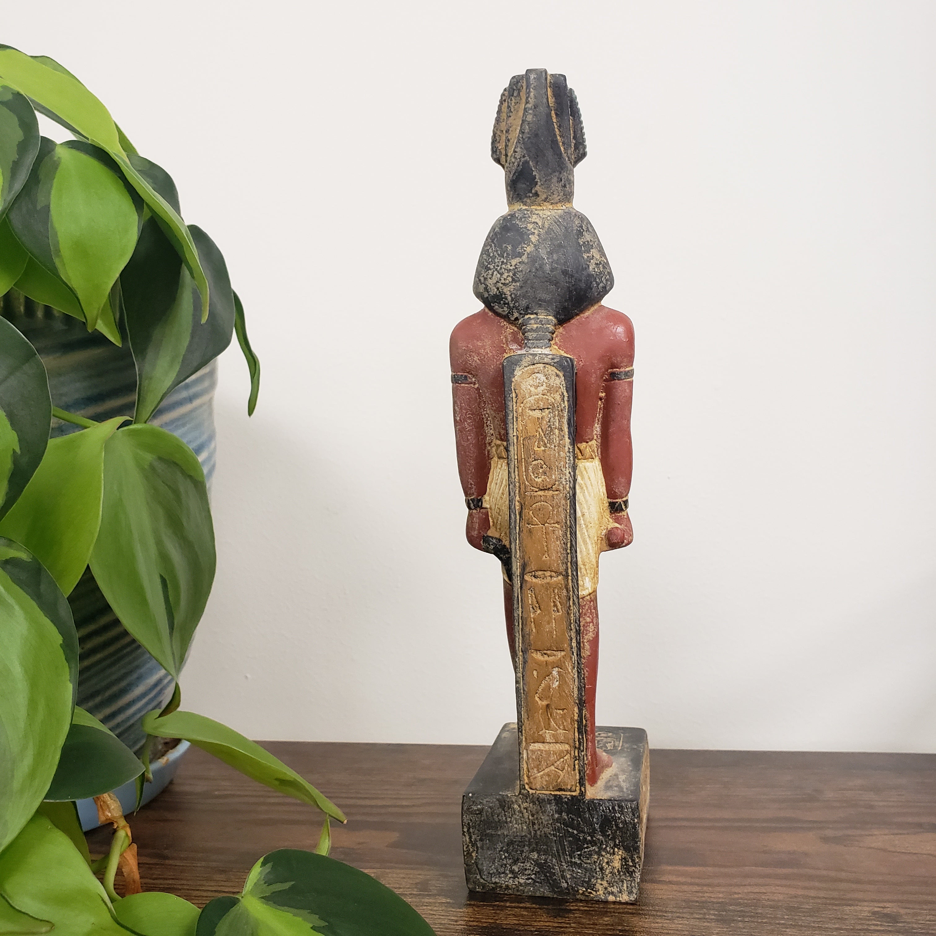 Egyptian Ramses statue with a red garment against a white wall with green plants in the foreground.