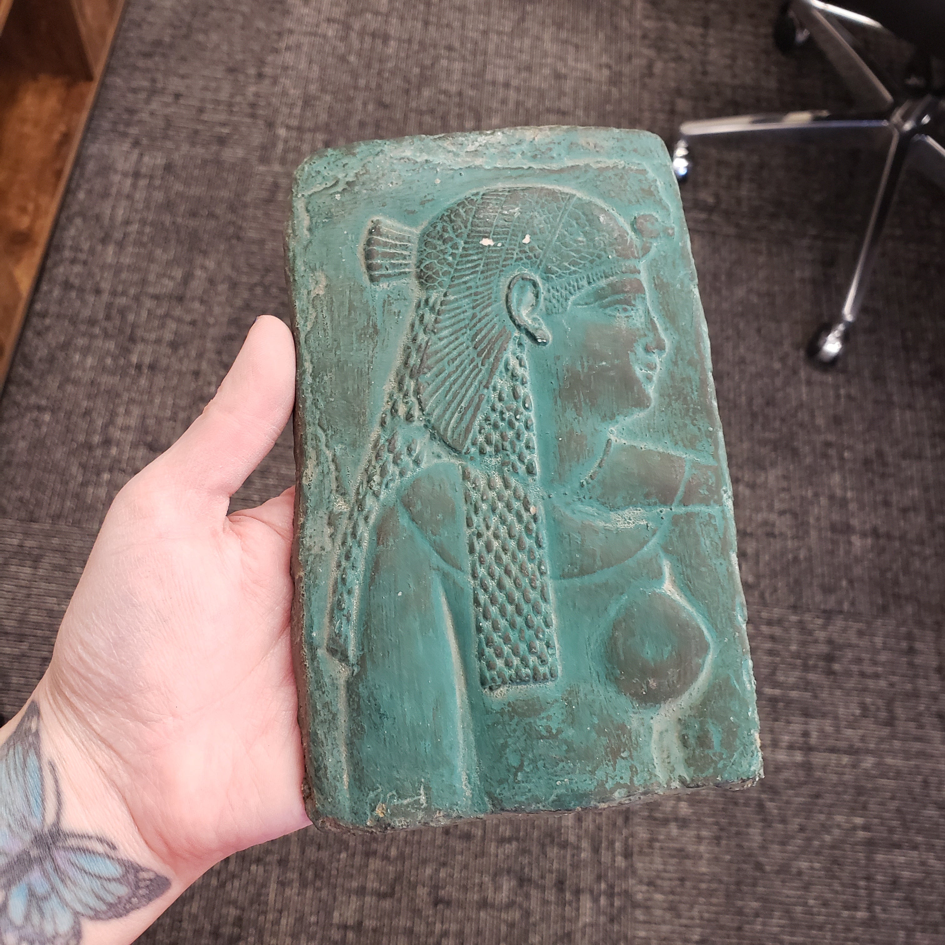Hand holding a greenish-brown Cleopatra wall hanging on a textured surface