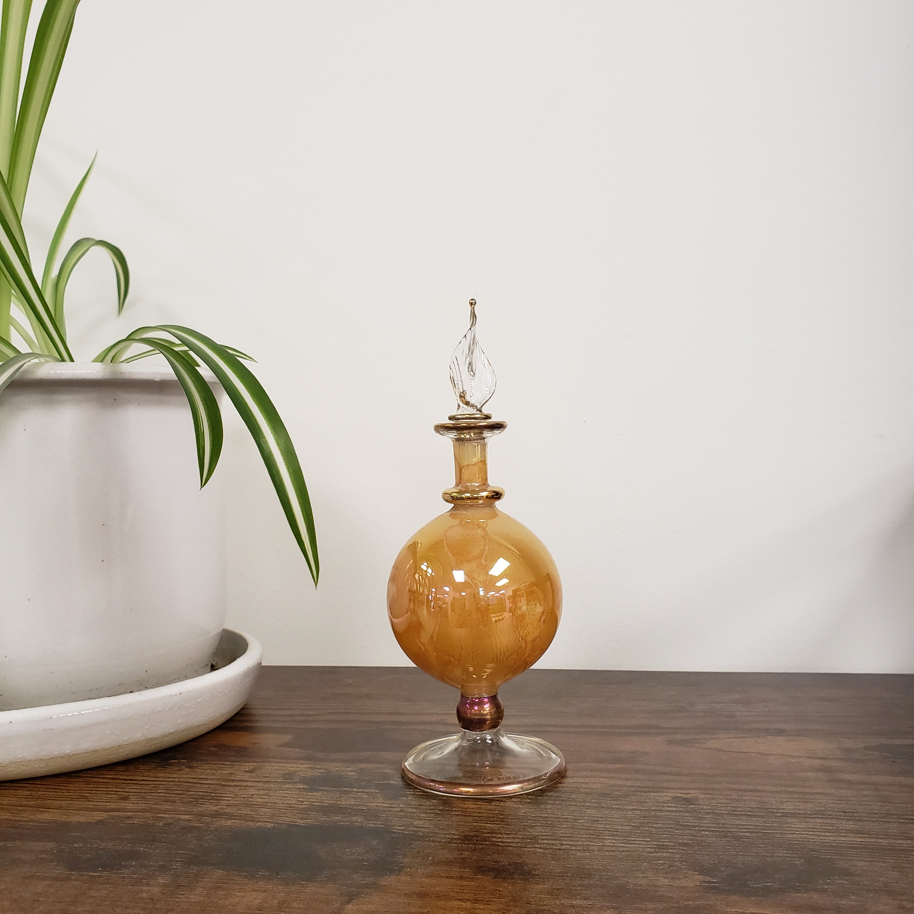 Decorative glass bottle with amber-colored bulb on a wooden surface with a plant in the foreground.