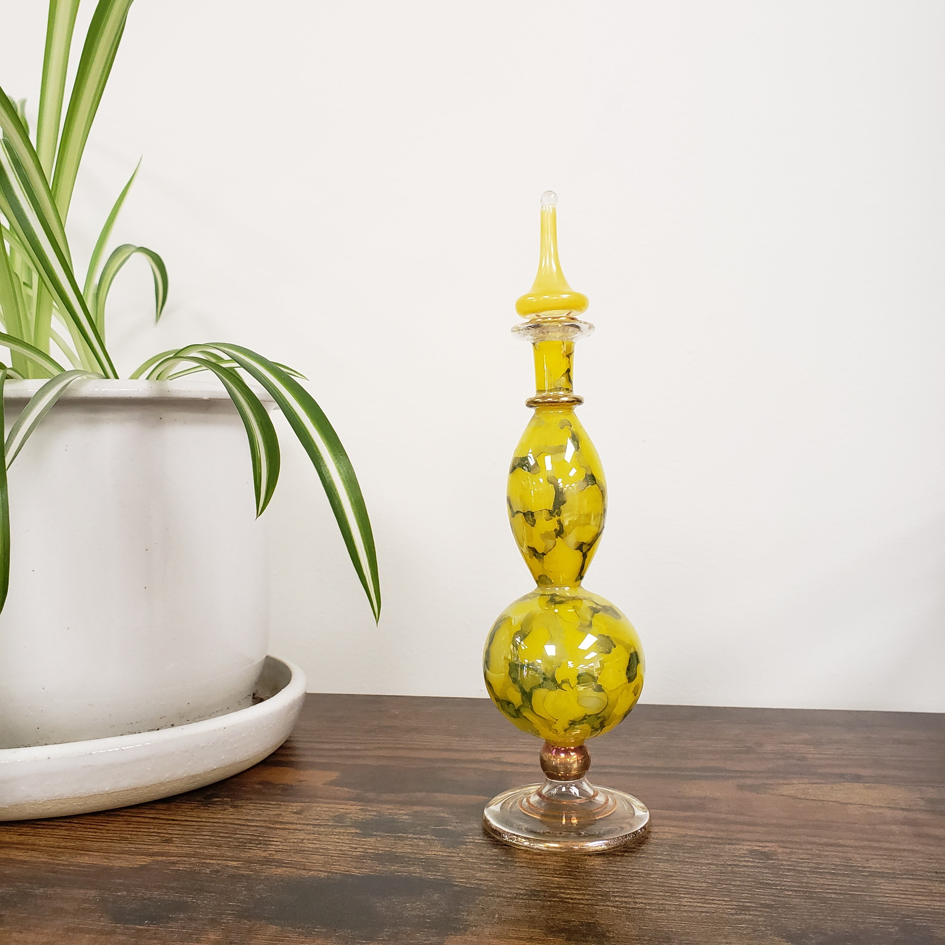 Decorative glass bottle with yellow and black pattern on a white surface with a plant and wooden surface in the background.