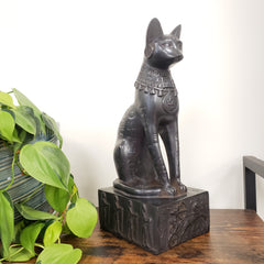 Decorative black cat sculpture on a wooden shelf with green plants below.
