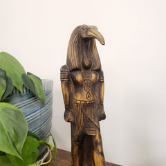 Wooden sculpture of a creature with a bird's head and human body, placed on a surface with green leaves in the foreground.