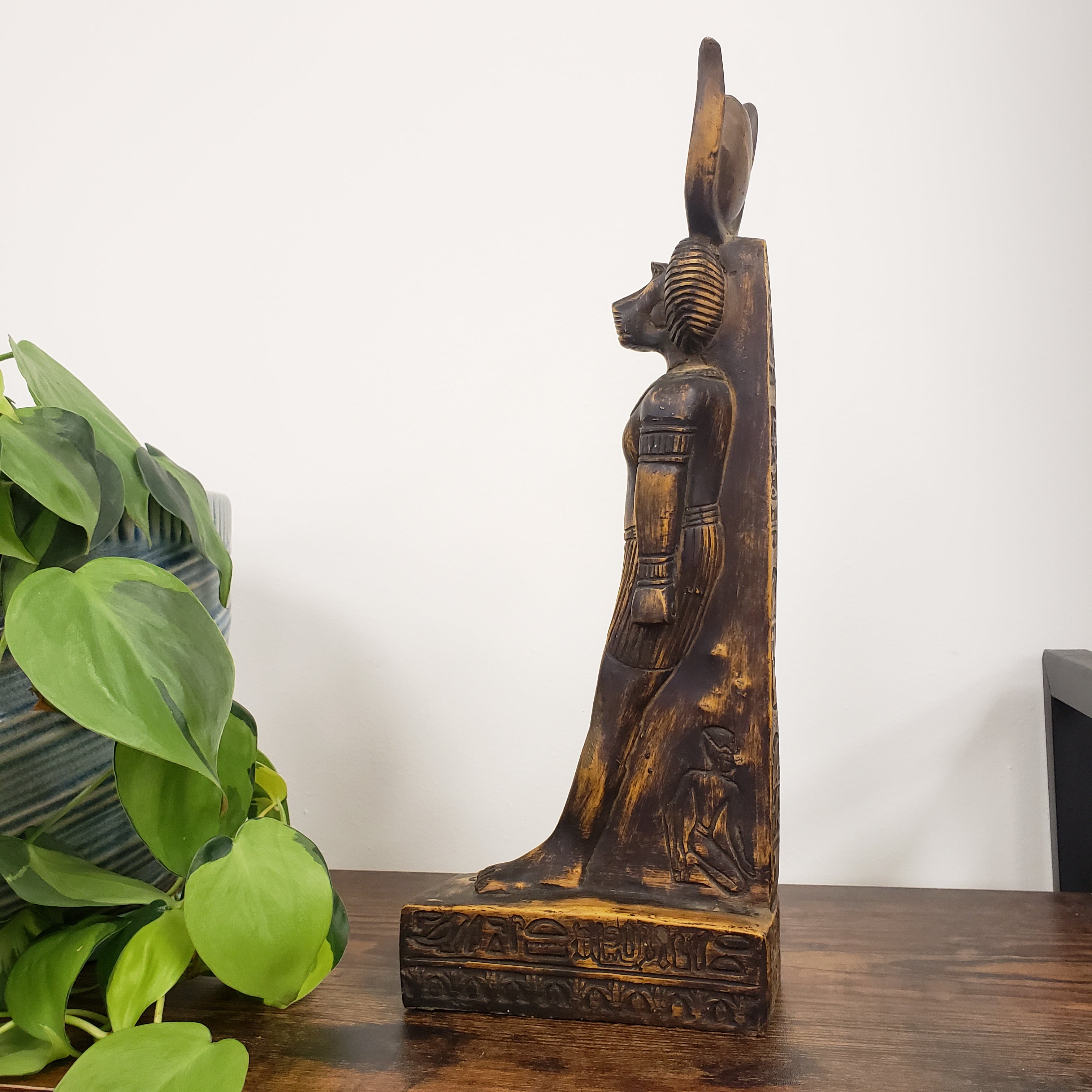 Decorative statue of the Egyptian god Babi with intricate carvings on a white wall with a plant in the foreground.