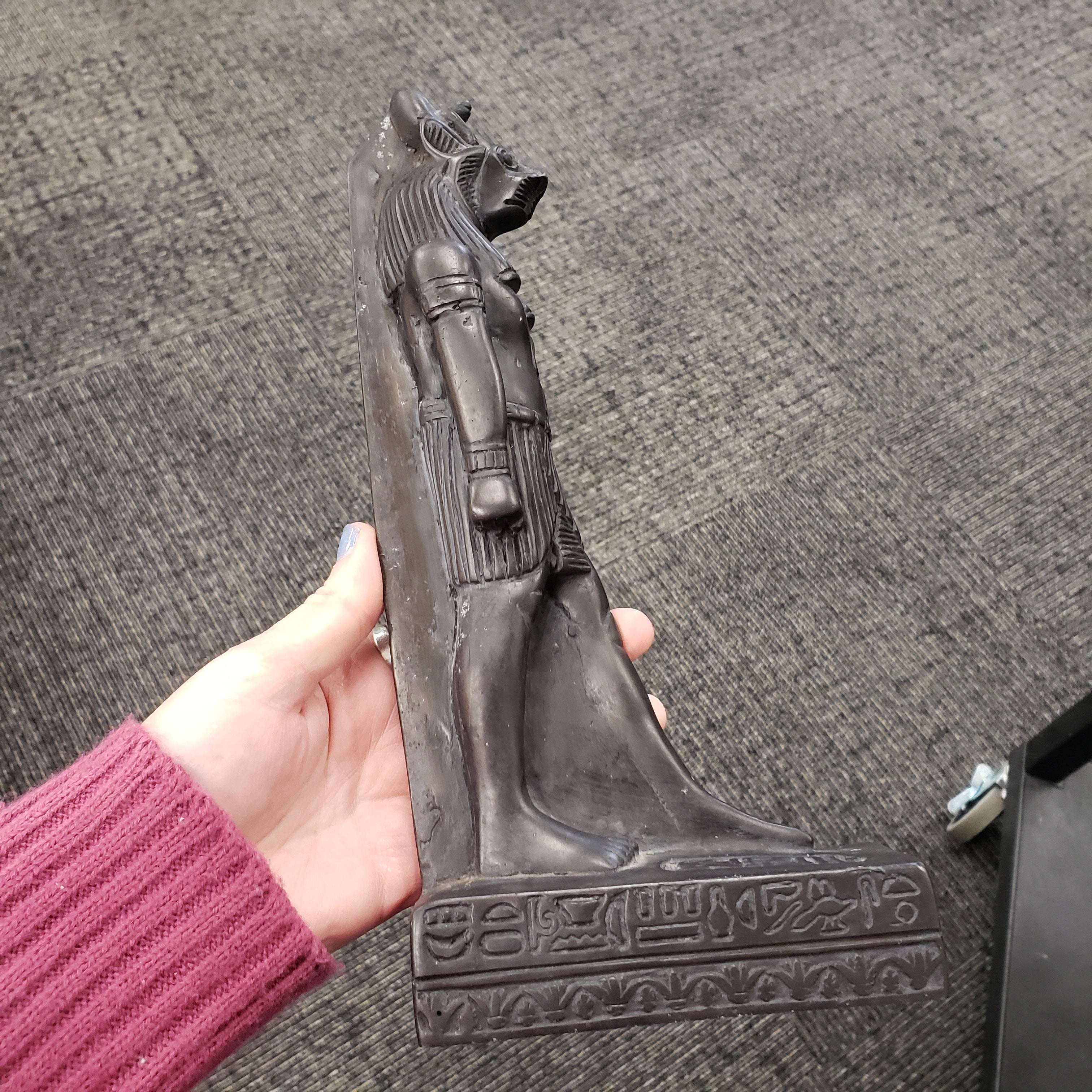 Hand holding a black Egyptian goddess Sekhmet statue with intricate designs on a gray carpeted floor.
