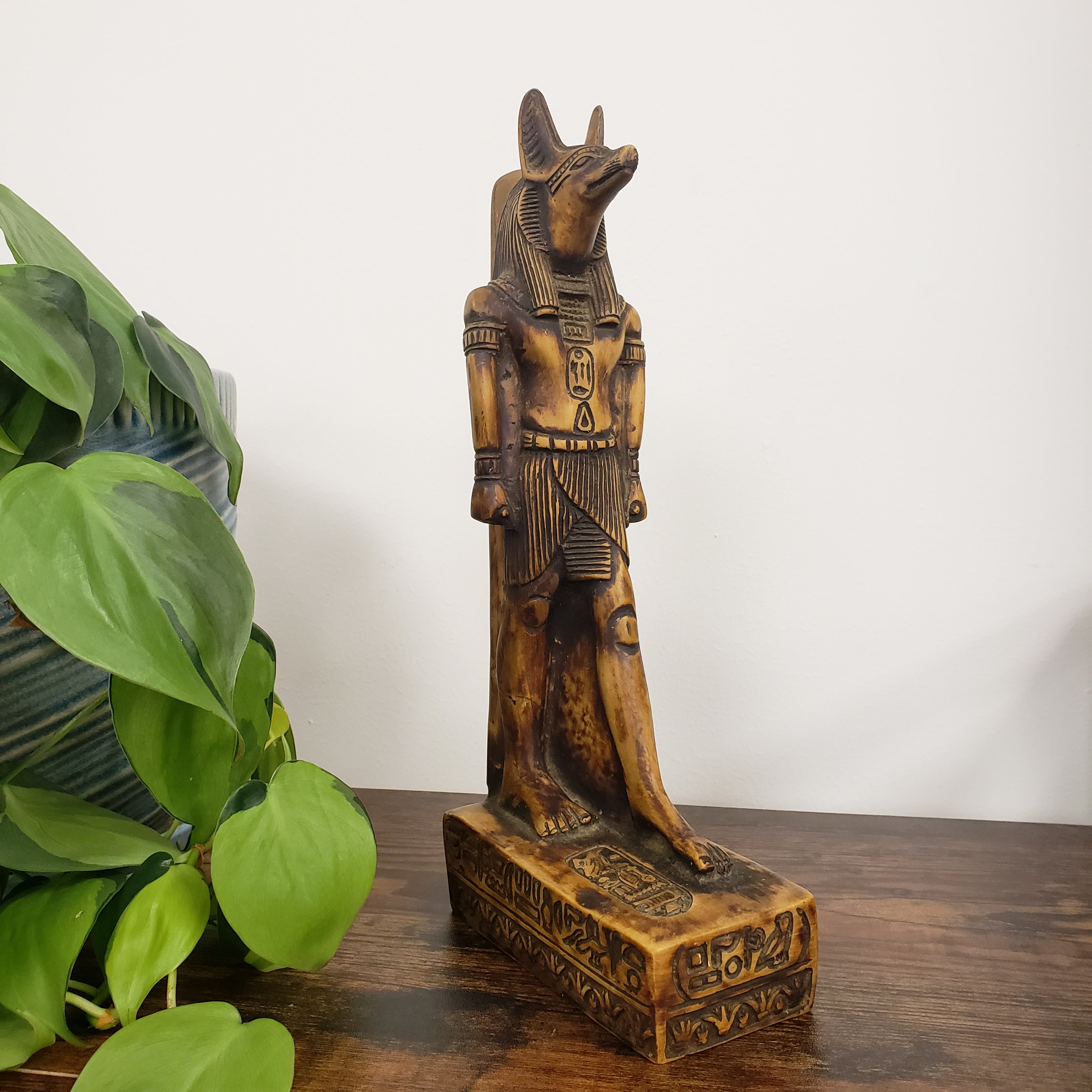 Decorative wooden sculpture of a figure with a block, placed against a white wall with a plant in the foreground.