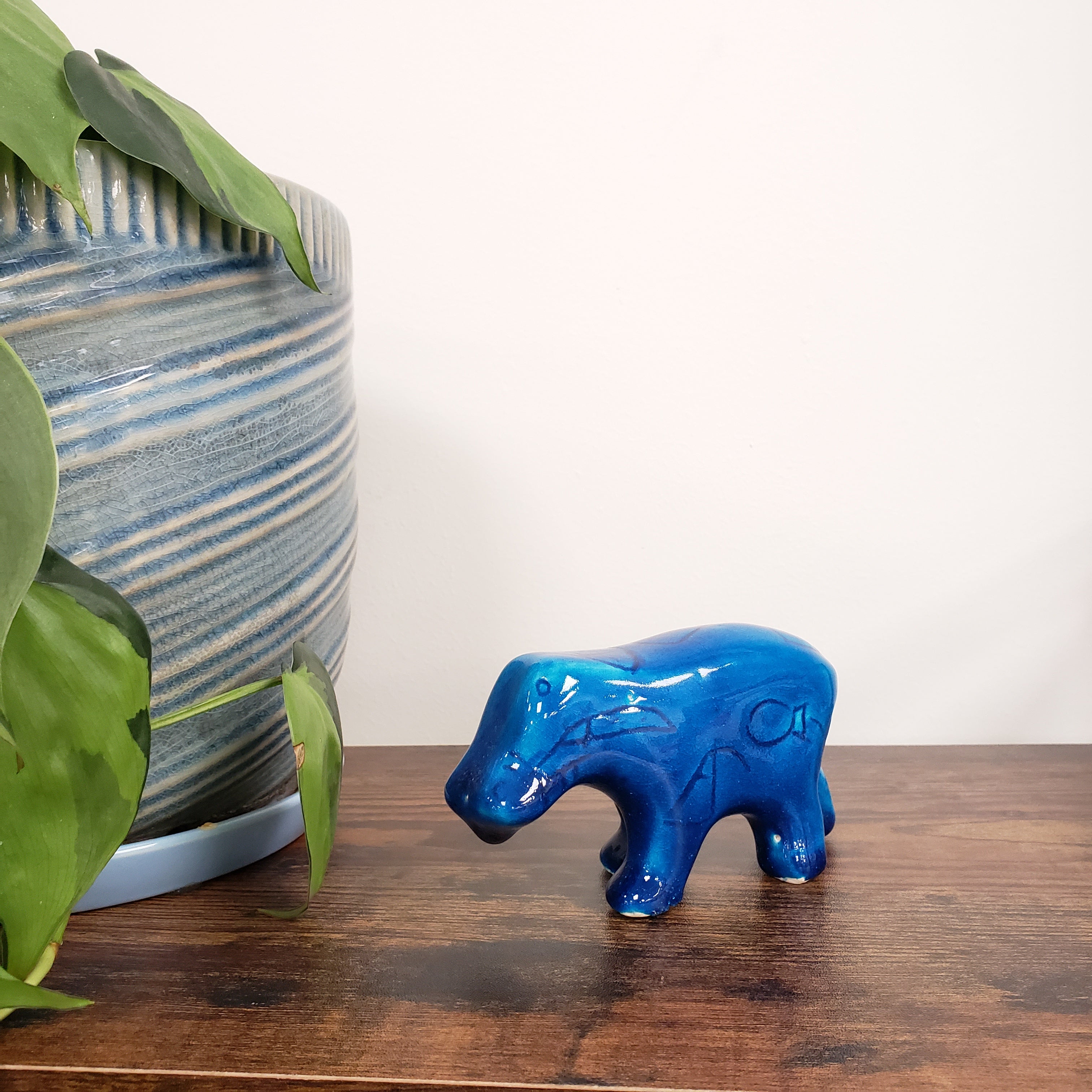 Blue ceramic hippo figurine on a wooden surface with green leaves and striped fabric in the background