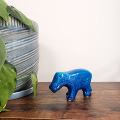 Blue ceramic hippo figurine on a wooden surface with green leaves and striped fabric in the background