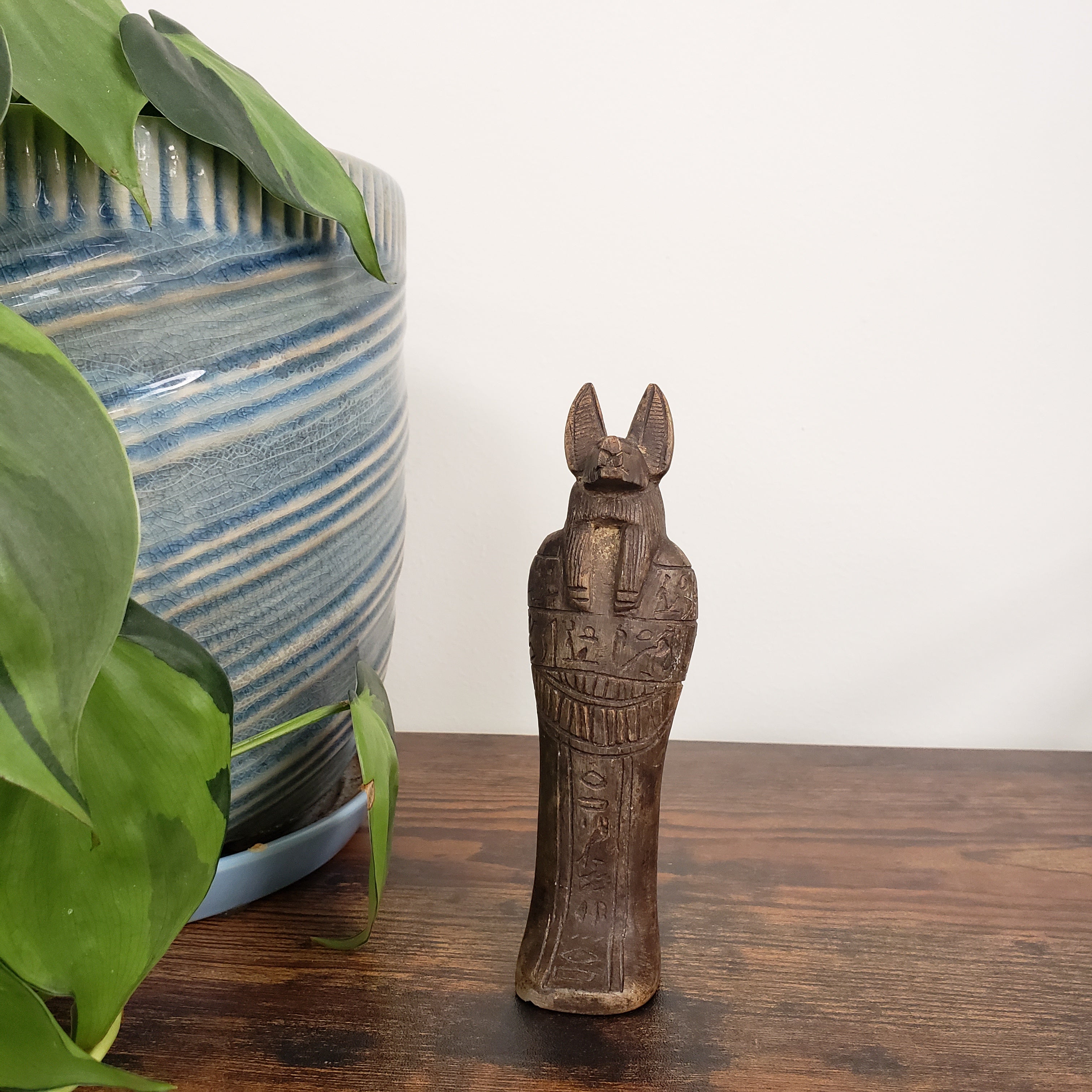 Wooden Anubis figure on a wooden surface with a plant and ceramic pot in the background.