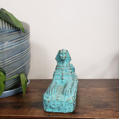 Decorative turquoise Egyptian Sphinx Statue on a wooden surface with a striped fabric and plant in the background.
