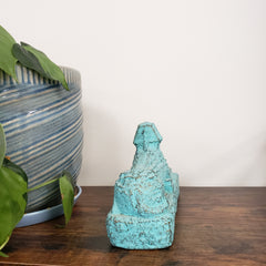 Turquoise Egyptian Sphinx Statue on a wooden shelf with a striped cushion and green leaves in the foreground.